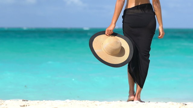 Lady on beach in black sarong standing on shore looking at ocean. Sexy female is enjoying wind on beach. Woman is holding sunhat during summer vacation by turquoise sea in the Caribbean.
