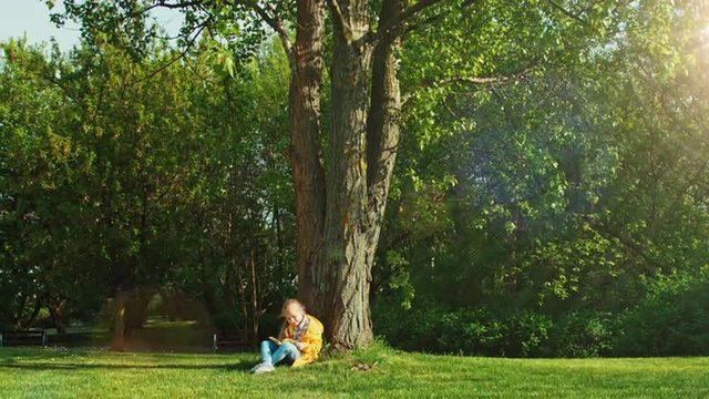 Child sitting under a tree and reading a book and looking at camera. Thumb up. Ok
