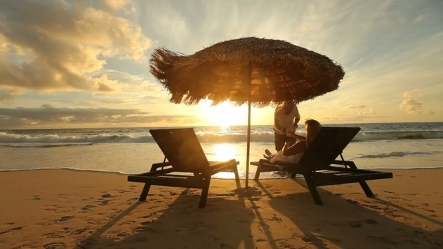 Man brings drink to woman sitting by beach at sunrise