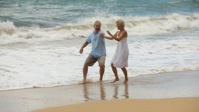 Senior couple run on tropical beach and play in surf