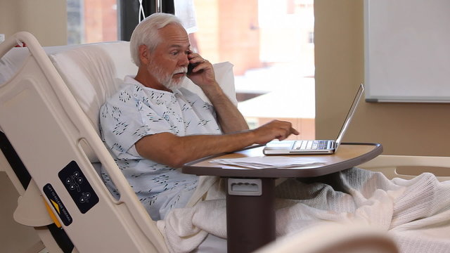Man in hospital bed uses phone and laptop comput