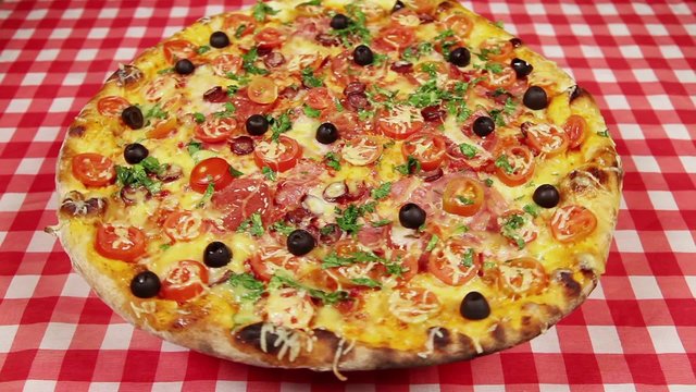 Low angle view video of rotating pizza with cheese, tomatoes and olives on the red tablecloth
