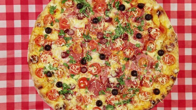 Front view video of rotating pizza with cheese, tomatoes and olives on the red tablecloth
