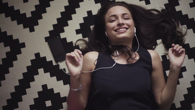 Attractive girl lies on the floor and listens to the music in her mobile device, earphones is plugged-in, she makes a lip sync and dances a bit.
