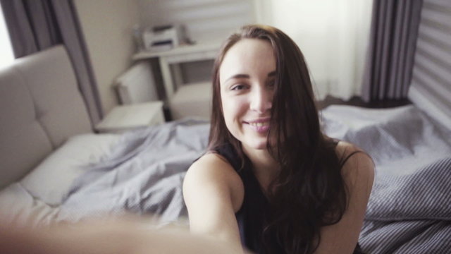 Slow motion footage of handsome cheerful girl enters a room and sit onto a bed,  selfie shot. 