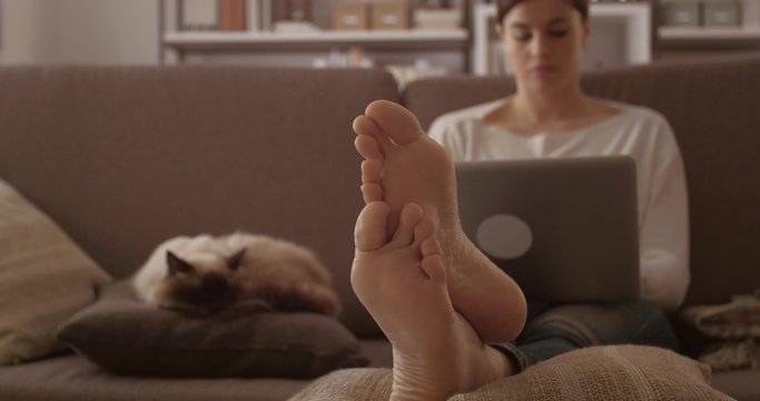 Young woman at home sitting on the sofa, she is working with a laptop and cuddling her beautiful birman cat sleeping next to her