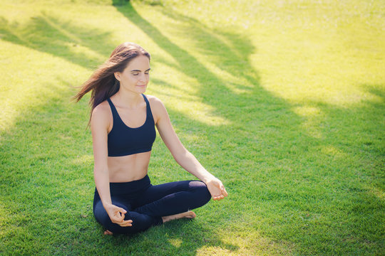 Молодая девушка занимается йогой. Девушка с длинными волосами сидит в позе лотоса на зеленом газоне.