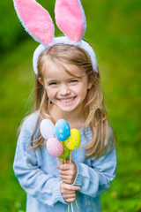Beautiful smiling little girl with long blond hair wearing pink rabbit or bunny ears and blue knitted dress and holding bunch of painted colour eggs on sunny day in spring park. Easter celebrations