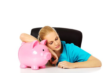 Young tired female doctor or nurse sitting behind the desk and holding a piggybank