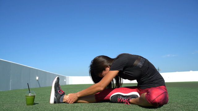 Fitness woman stretching hamstring - glute exercising toe-touch stretches. Sporty young athlete in activewear doing a one leg stretch on grass in sunny outdoor gym with green smoothie cup on the side.