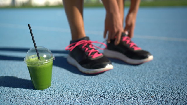 Female athlete runner getting ready for run race with green smoothie breakfast plastic cup tying running shoes on blue outdoor athletic track. Feet closeup.