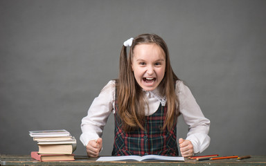 Ребенок в школьной форме  злится, кричит и стучит кулаками по столу с книгами на сером фоне 