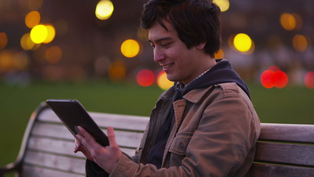 Young man sits on park bench using digital tablet