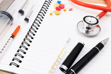 Red stethoscope, syringes, pen, and many colorful pills on blank notepad