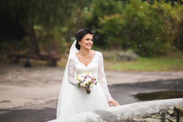 Beautiful brunette bride in elegant white dress holding bouquet posing neat trees 
