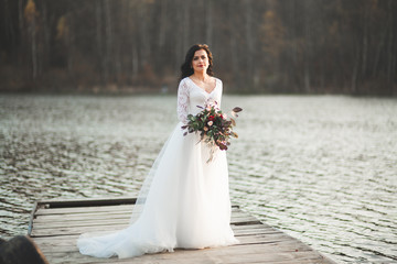 Fototapeta na wymiar Gorgeous brunette bride in elegant dress holding bouquet posing near forest and lake