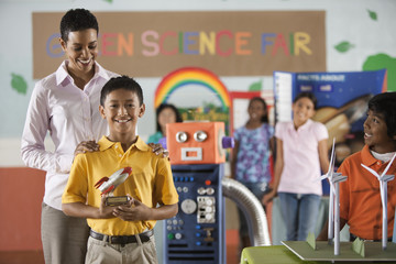 A boy holding a rocket trophy standing in front of the class, with an adult, winning a trophy at the Green Science Fair,
