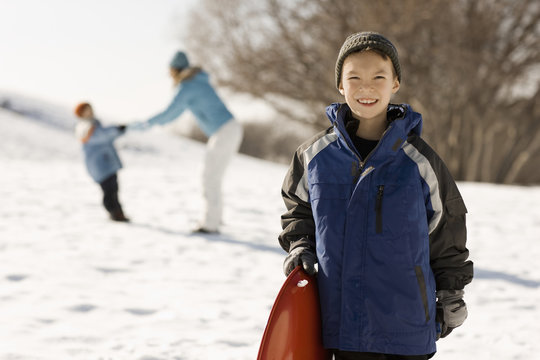 A boy holding a sledge in snow, and a mother and child behind him, 
