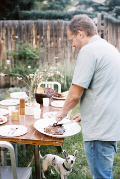 A man carving meat at a family meal in a garden, being watched by a small dog under the table,