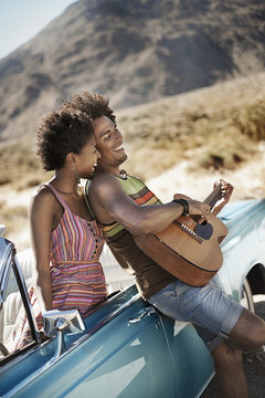 A young couple standing by a pale blue convertible on the open road, the man playing a guitar, 