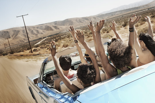 A group of friends in a pale blue convertible on the open road, driving across a dry flat plain surrounded by mountains, 