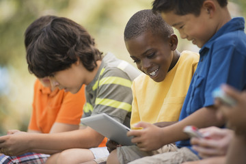 A row of children sitting outdoors in summer using tablets and handheld games, 