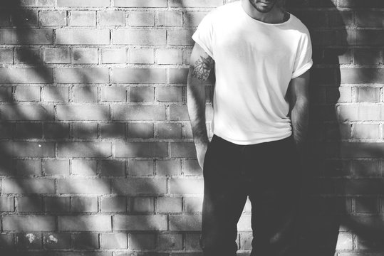 Photo bearded man with tattoo wearing blank white tshirt and black jeans. Bricks wall background. Horizontal mockup, blackwhite