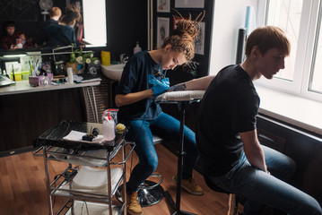 Tattoo girl with dreadlocks on his head in the stuffing sleeve tattoo in black and white. Master works in a professional salon on a white mat and in sterile blue gloves.