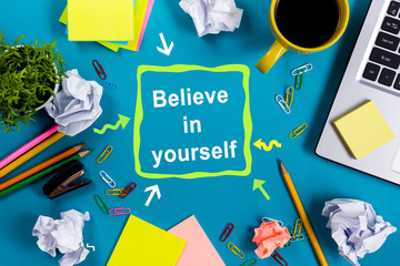 Believe in yourself. Office table desk with supplies, white blank note pad, cup, pen, pc, crumpled paper, flower on wooden background. Top view
