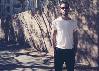 Bearded man with tattoo wearing blank white tshirt and black sunglasses. City garden wall...