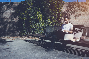 Photo bearded man wearing white tshirt sitting city park bench and reading book. Studying at the University, working new project. Books, laptop, backpack bench. Horizontal