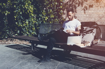 Fototapeta premium Photo bearded man wearing white tshirt sitting city park bench and reading book. Studying at the University, working new project. Books, laptop, backpack bench. Horizontal mockup