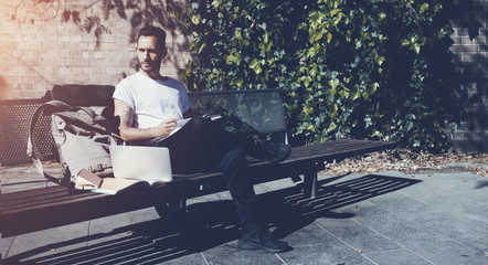 Fototapeta premium Bearded man wearing white tshirt sitting city park bench and reading book. Studying at the University, working new project. Books, laptop, backpack bench. Horizontal 