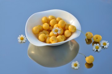  yellow cherry, camomile flowers in a white plate on a mirrored surface