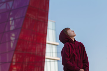 Urban portrait of teenage boy wearing maroon hooded sweatshirt  at modern glass skyscraper city background 
