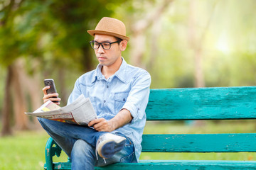 Asian young Man with hat sitting on a wooden bench and reading a newspaper and checking message by smartphone screen with a serious expression in a park