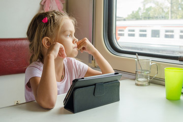 Girl thoughtfully looked out the window while sitting with a tablet on a train - obrazy, fototapety, plakaty