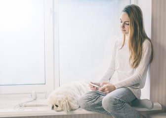 Beautiful girl with her dog sitting on the windowsill and use tablet pc at home.