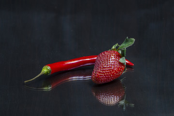 Red hot chili pepper and a  strawberry are on a mirror black wooden table