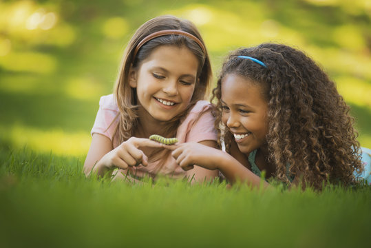 Two girls lying on the grass, one holding a green caterpillar on her finger, 