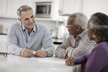 Three people at a table, a senior couple seated with a man using paper and pen to give them information, 