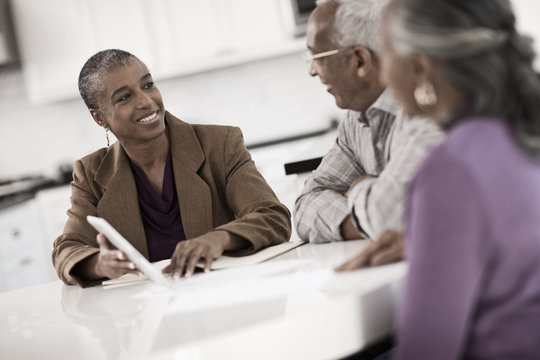 Three people at a table, a senior couple seated with a woman showing them a digital tablet screen, 