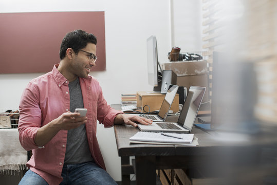 A man in a home office with a desk with two laptops, checking information on the screen, holding a smart phone,