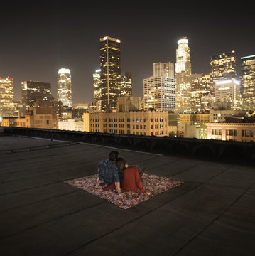 A couple sitting on a rug on a rooftop overlooking a city lit up at night, 