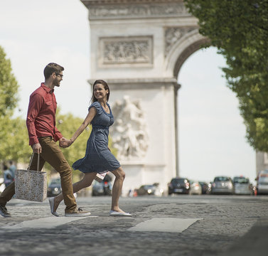 A couple hand in hand carrying shopping bags and crossing the road by a historic monument in the heart of a European city, 