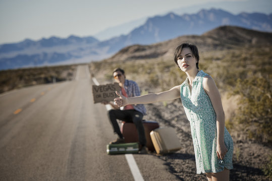 A young couple, man and woman, on a tarmac road in the desert hitchhiking, with a sign saying Vegas or Bust,
