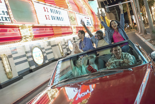 A group of friends in a red open top convertible classic car celebrating with waving arms as they drive through a city lit with neon signs, 
