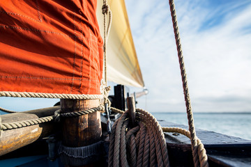 rope wound on a wooden cleat fixed on the hull of a rigging vintage sailing boat with a beige jib and an ocher sail filled by the wind at the blur background during a sunny sea trip in brittany - obrazy, fototapety, plakaty