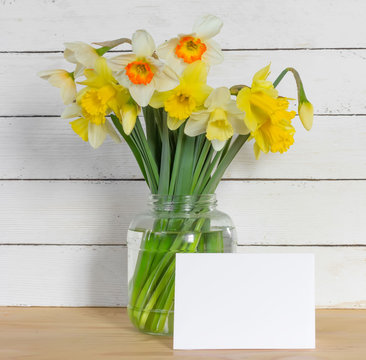 Bouquet of daffodilsin a glass jar with a blank greeting card on a wooden table in the background of the white wooden wall with copy space. Provence style. Square photo