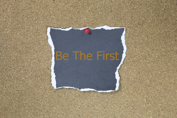 The phrase Be The First in a black sticky note posted to a cork notice board.